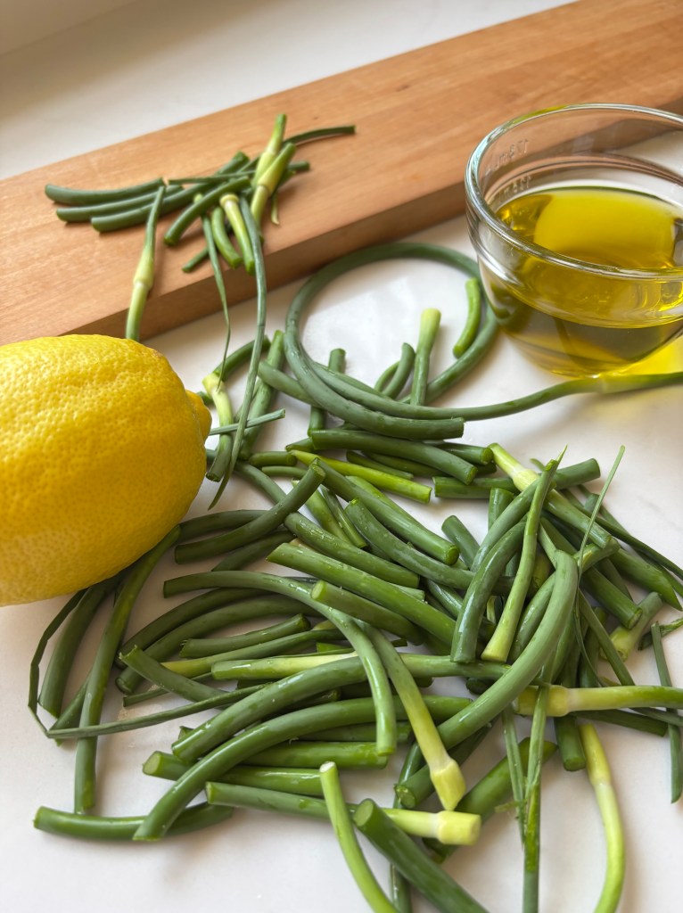 The ingredients for garlic scape pesto on a white countertop with a long brown cutting board: lemon, oil, garlic scapes.