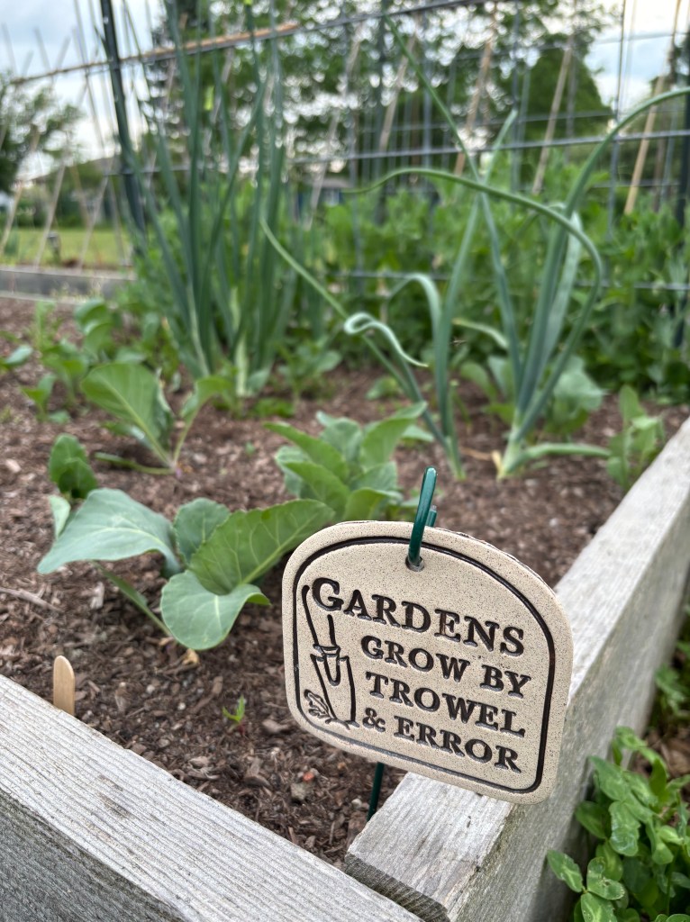 A small sign stuck in a raised garden bed that reads "Gardens grow by trowel and error".